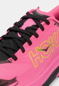 Lys rosa joggesko med svarte snørebånd, med en teksturert overdel, polstret såle, og "HOKA"-logo i gult med svart aksent.