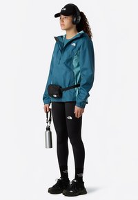 Turkoosi kevyt vedenpitävä takki vetoketjulla, musta vyölaukku, mustat leggingsit, mustat lenkkarit ja hopea vesipullo. Mukana kuulokkeet ja lippalakki.