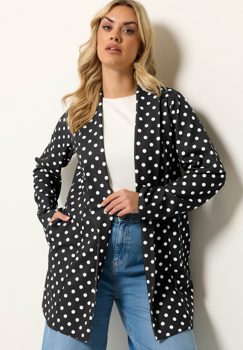 Kvinde med langt blondt hår iført sort blazer med hvide prikker, hvid skjorte og blå jeans, står foran en ensfarvet lys baggrund.