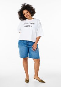 White cropped t-shirt with black text "TROUVEZ VOTRE HARMONIE," paired with blue denim bermuda shorts and olive green pointed flats.