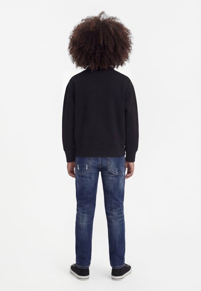 Παιδί με σγουρά μαλλιά που στέκεται, φοράει μαύρο μακρυμάνικο πάνω μέρος, μπλε τζιν και μαύρα αθλητικά παπούτσια, βλέποντας προς τα πίσω σε λευκό φόντο.