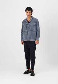Chaqueta de pana gris con dos bolsillos frontales, cierre de botones y cuello, llevada sobre una camisa burdeos y pantalones negros.