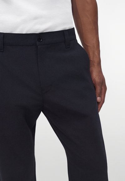 Personne portant un pantalon noir avec fermeture par bouton et fermeture éclair, poche latérale visible, et une chemise blanche rentrée, sur un fond uni.