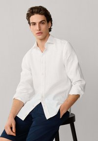 Joven con cabello rizado castaño, vestido con una camisa blanca de botones con las mangas remangadas y pantalones cortos azul marino, sentado en un taburete negro.