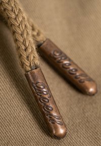 Primer plano de una cuerda trenzada beige con puntas metálicas de color cobre estampadas con texto en cursiva sobre un fondo de tela texturizada.