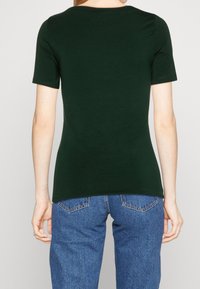 Camiseta de manga corta verde oscuro con cuello redondo, textura suave y silueta ajustada, combinada con jeans de color azul claro.