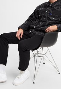 Una persona è seduta su una sedia moderna, indossando una giacca nera a fantasia, pantaloni neri e sneakers bianche, mentre riposa casualmente il braccio sulla sedia.