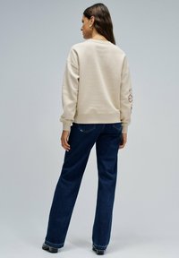 Mujer con cabello largo que lleva una sudadera beige con diseño en las mangas, jeans oscuros y zapatos negros, de pie de espaldas contra un fondo liso.