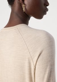 Ljust beige tröja med rund hals, mjuk textur och synliga sömnadsdetaljer, bärs med en enkel guldförsedd örhänge som framhäver designen.