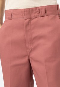 Růžové kalhoty na míru mají hladkou látku s jemným leskem, rovný přední díl a boční kapsy. Pas je strukturovaný pro čistý střih.