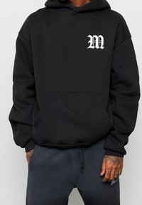 Sudadera negra con bolsillo frontal y logo blanco "M". Fabricada en un tejido suave, con un corte holgado y puños acanalados.
