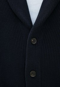 Primo piano di un cardigan blu navy lavorato a maglia con due bottoni scuri sopra una camicia bianca.