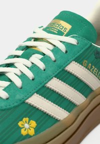 Πράσινο σουέντ αθλητικό παπούτσι με λευκές δερμάτινες ρίγες, με κίτρινο σχέδιο λουλουδιού και χρυσό λογότυπο. Υφή σόλας από καουτσούκ. Κλασική αισθητική.
