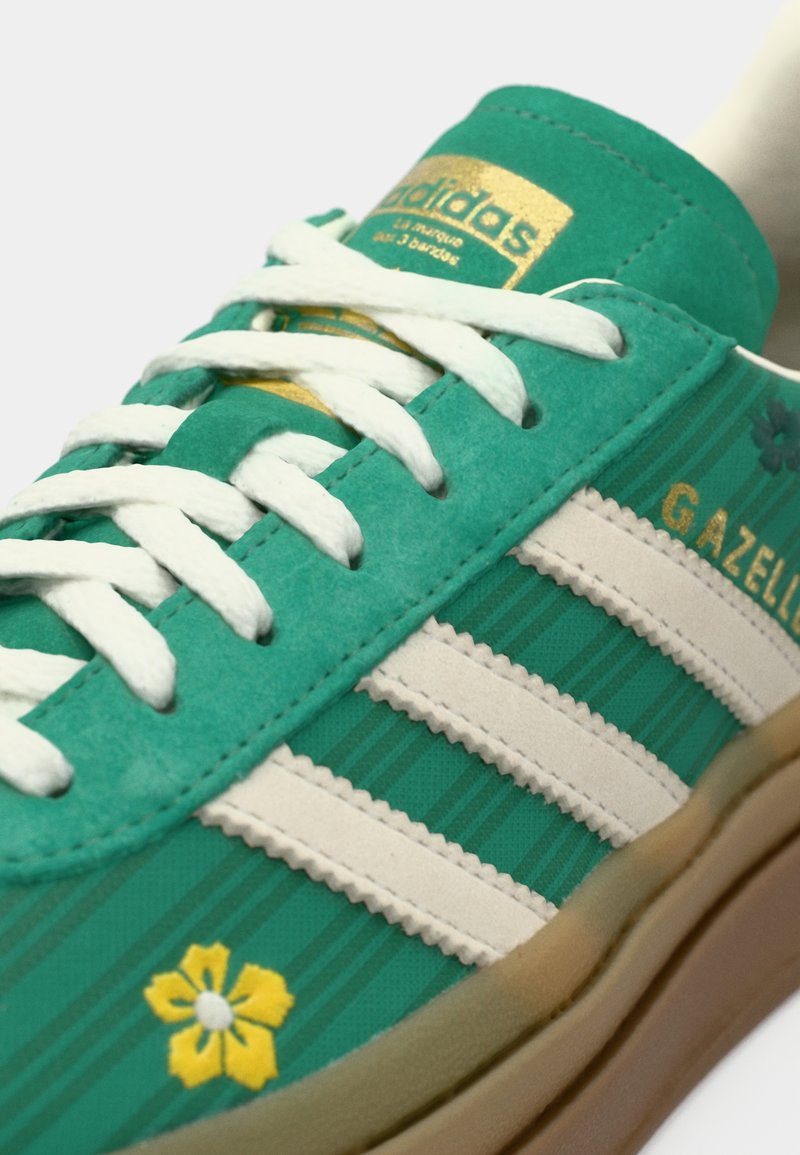 Zapatilla deportiva de ante verde con franjas de cuero blanco, con un diseño de flor amarilla y marca dorada. Suela de goma texturizada. Estética clásica.