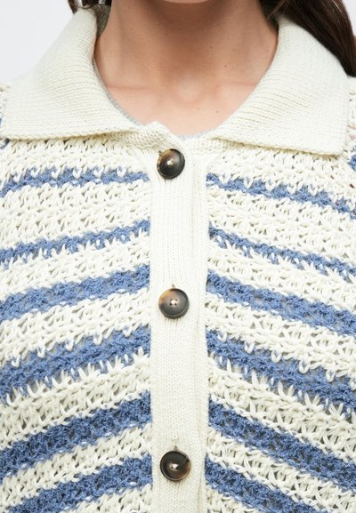 Κοντινό πλάνο πλεκτής ζακέτας με λευκές και μπλε διαγώνιες ρίγες, φαρδιά γιακά και τέσσερα σκοτεινά κουμπιά μπροστά.