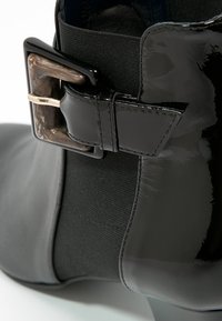 Bottine noire brillante avec une boucle carrée en écaille de tortue et un panneau latéral élastique sur un talon bas.