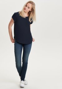 Navy short-sleeved top with rounded hem, paired with dark blue skinny jeans. Model wears white sneakers. Light background.