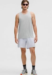 Homme portant un débardeur d'entraînement sans manches gris clair, un short assorti, des chaussettes noires à motifs, des chaussures de course grises et des lunettes de soleil foncées, se tenant sur un fond blanc.