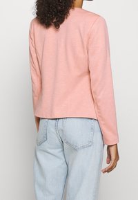 Vaaleanpunainen, pitkähihaista takkia, jossa on sileä pinta, slim fit -malli ja pyöreä helma. Takapuolelta näkyy ompelut yksityiskohtia ja vaaleat farkut.