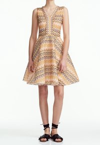 Abito senza maniche a V con gonna svasata in un motivo zigzag giallo, rosa e nero, indossato con sandali piatti neri allacciati.