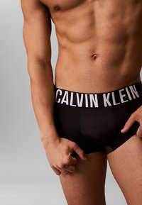 Zwarte boxer briefs met een witte Calvin Klein logo tailleband, met een strakke pasvorm en een zachte textuur, deels verborgen door een hand.