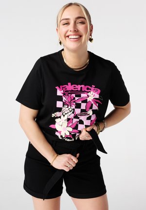 Glimlachende vrouw in een zwarte korte broek en een zwart t-shirt met een roze bloem- en geruit patroon met het label "Valencia." Ze houdt een zwarte riem vast.