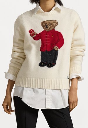 Pull avec un ours en peluche marron tricoté portant une veste rouge tenant un livre rouge, porté sur une chemise blanche et un pantalon noir.
