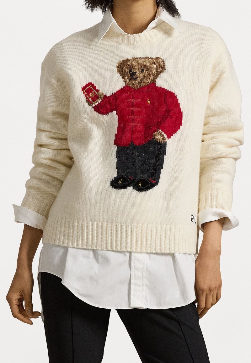 Pull avec un ours en peluche marron tricoté portant une veste rouge tenant un livre rouge, porté sur une chemise blanche et un pantalon noir.