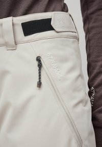 Lichtbeige broek gemaakt van zachte stof, met een ritssluiting op de zak, zwarte accenten aan de tailleband en een geborduurd logodetail.