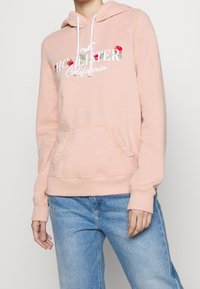 Różowa bluza z kapturem z przednią kieszenią, ozdobiona kwiatowym motywem i napisem. Materiał wygląda na miękki i teksturowany, z ściągaczami na mankietach i u dołu.