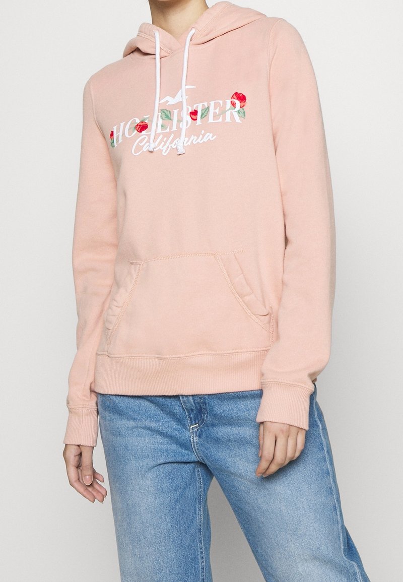 Różowa bluza z kapturem z przednią kieszenią, ozdobiona kwiatowym motywem i napisem. Materiał wygląda na miękki i teksturowany, z ściągaczami na mankietach i u dołu.