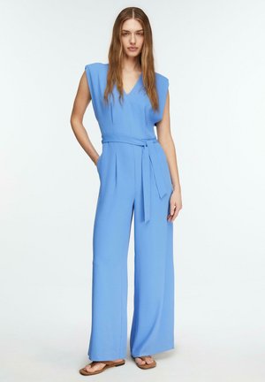 Kobieta stojąca w jasnoniebieskim, bezrękawnym kombinezonie z paskiem, o szerokich nogawkach, w beżowych, płaskich sandałach na tle białego tła.