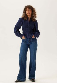 Vrouw met krullend haar die een marineblauwe blouse met geplooide details draagt, een high-waisted jeans met wijd uitlopende pijpen en zwarte ballerina's, staand met handen in de zakken.