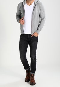 Grauer Reißverschluss-Hoodie mit einem weißen V-Ausschnitt-T-Shirt, dunklen Jeans und braunen Schnürstiefeln. Lässiges Design mit bequemer Passform und steigendem Kragen.