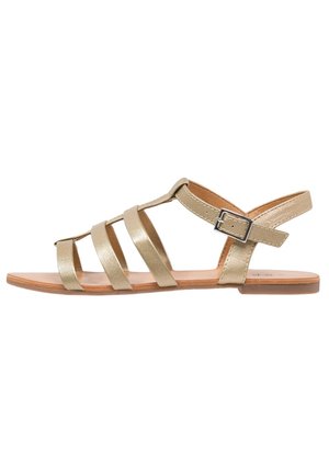Flad gylden sandal med fire frontremme, en ankelrem med spænde og en tynd sål på hvid baggrund.