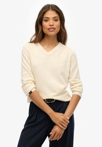 Creme V-hals trui gemaakt van zacht materiaal, met omgeslagen mouwen en een slanke pasvorm. Gecombineerd met marineblauwe gestreepte broeken voor contrast.