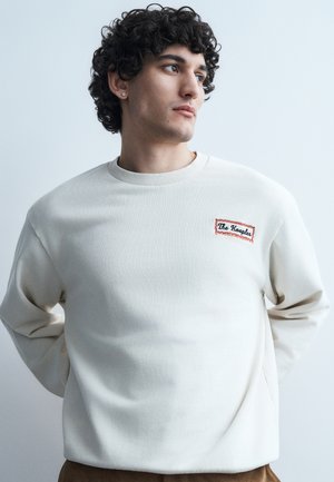 Νεαρός με σγουρά μαλλιά που φοράει λευκό φούτερ με μπαλώματα "The Kooples", κοιτάζει προς το πλάι με φόντο ένα απλό φωτεινό τοίχο.