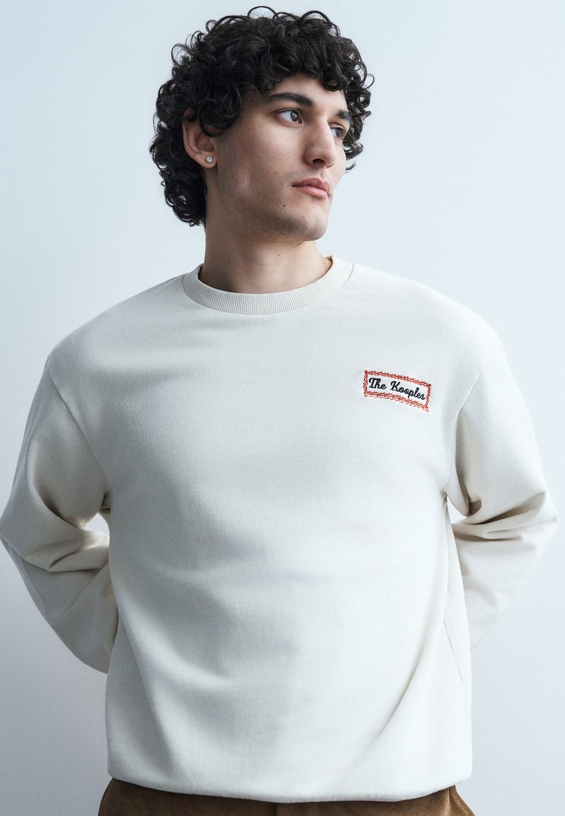 Νεαρός με σγουρά μαλλιά που φοράει λευκό φούτερ με μπαλώματα "The Kooples", κοιτάζει προς το πλάι με φόντο ένα απλό φωτεινό τοίχο.