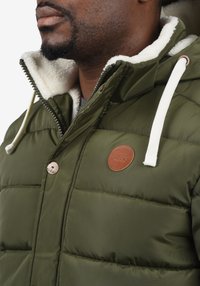 Olive green padded jacket with white faux shearling lining, brown logo patch, and drawstring hood. Features metallic zipper and snap buttons.