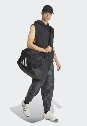 Borsa duffel nera con tre strisce bianche, materiale in tessuto, design spazioso, indossata da una persona con una felpa smanicata e pantaloni jogger fantasia.