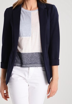 Vrouw draagt een marineblauwe blazer met opgerolde mouwen over een kleurvlak-T-shirt en witte broek, staand tegen een effen achtergrond.