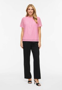 Blouse en dentelle rose à manches courtes, col arrondi et ourlet festonné, associée à un pantalon noir à jambes droites et des sandales noires à talons.