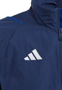 Marineblaue Zip-Jacke mit hohem Kragen, blauen Akzentnähten und einem weißen gestickten Logo auf der linken Brust. Glatte Stofftextur.