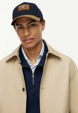 Jonge man met een beige jas, marineblauwe trui met een kwart rits, lichtblauw gestreept overhemd en een marineblauwe pet met een rechthoekige bruine patch.