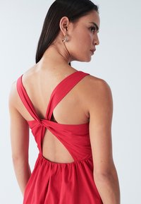 Rotes Kleid mit breiten Trägern, das ein gedrehtes Rückendesign, eine taillierte Passform und eine offene Rückseite zeigt; glatte Textur und leichter Stoff.