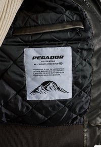 Mustast järjestatud vooder beeži kudumisega kaeluse ning valge sildiga, millel on kiri "PEGADOR OUTERWEAR" ja mägigraafika.