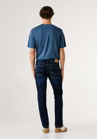 Modrá bavlnená tričko a tmavomodré džíny. Džíny majú kontrastné švy a zadné vrecká. Model nosí béžové topánky.