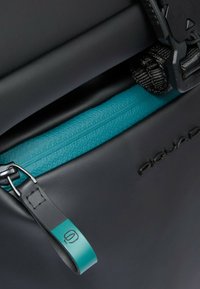 Zwarte waterdichte tas met een turkooise ritssluiting, gestructureerde stof en een matte afwerking. Voorzien van een zwarte band met hardware en merkdetail.