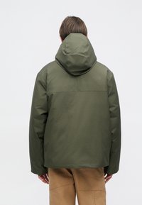 Maslinasto zelena jakna s kapuljačom, s mat završetkom i suptilnim oznakama na leđima. Labav kroj s ravnim rubom.
