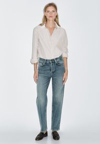 Gestreifte Hemd mit Knöpfen in Weiß und Pastellfarben, kombiniert mit hellblauen Jeans. Modell trägt dunkle Schuhe und steht vor einem schlichten Hintergrund.